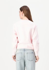 Sweatshirt en coton rose à manches longues, avec des poignets côtelés et un détail de couture au dos, associé à un jean bleu clair visible à la taille.
