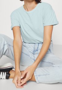 T-shirt di cotone azzurro chiaro con maniche corte arrotolate, abbinata a jeans di denim chiaro. Sneakers bianche con dettagli neri e lacci.