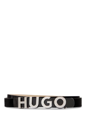 Musta kiiltävältä nahalta valmistettu vyö, jossa on suuret hopeiset metallikirjaimet muodostamassa lukkoa "HUGO" valkoisella taustalla.