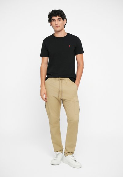 Juoda medvilnė marškinėliai su maža raudona logo, derinami su smėlio spalvos kargo kelnėmis su šoninėmis kišenėmis ir raišteliu, užbaigti baltais sportbačiais.