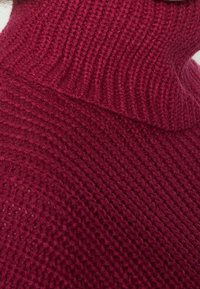 Sweter z dzianiny z golfem w głębokim odcieniu burgundu, z teksturowanym, prążkowanym wzorem. Widok z bliska podkreśla miękkość i grubość materiału.