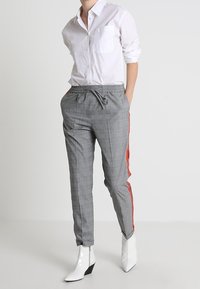 Šedé kostkované kalhoty s červenými bočními pruhy, elastickým pasem a volným střihem, kombinované s bílou košilí s knoflíky a bílými kotníkovými botami.