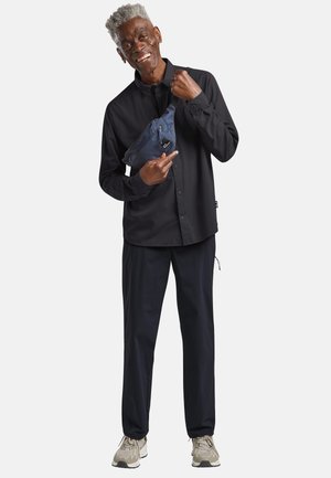 Ein lächelnder älterer Mann in schwarzem Hemd und Hose zeigt auf eine marineblaue Umhängetasche, die quer über seiner Brust getragen wird, vor einem schlichten Hintergrund stehend.