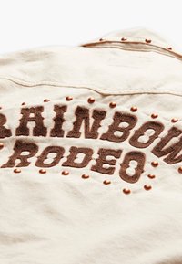 Off-white denimová košile s výšivkou textu &quot;RAINBOW RODEO&quot; v tmavě hnědé barvě, zvýrazněná měděnými cvočky podél límce.