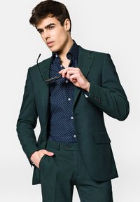 Ciemnozielona, dopasowana marynarka i spodnie z granatową koszulą w kropki; trzymający okulary przeciwsłoneczne, z klapami w kształcie notcha i kontrastowymi guzikami.