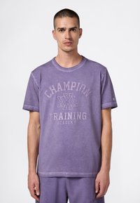 Fioletowa bawełniana koszulka z okrągłym dekoltem, krótkimi rękawami oraz nadrukiem "CHAMPION XXL TRAINING ACADEMY" z przodu.