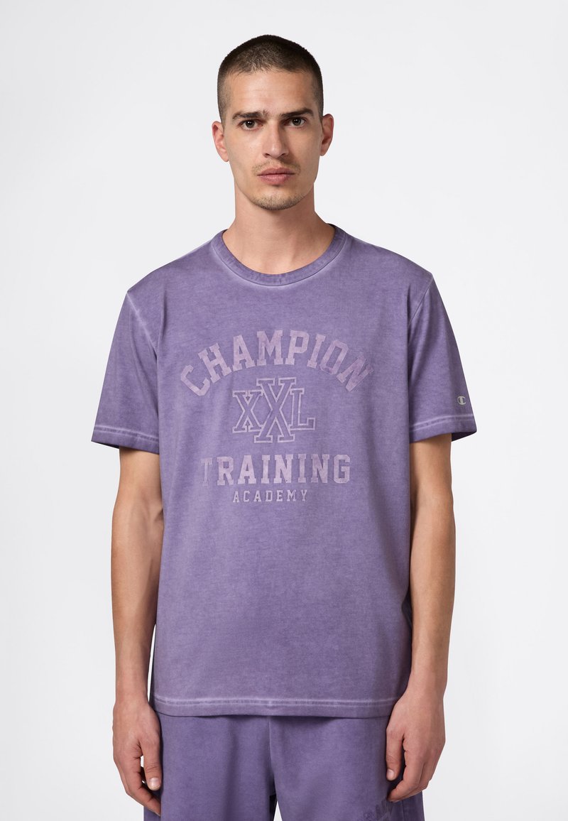 Fioletowa bawełniana koszulka z okrągłym dekoltem, krótkimi rękawami oraz nadrukiem "CHAMPION XXL TRAINING ACADEMY" z przodu.