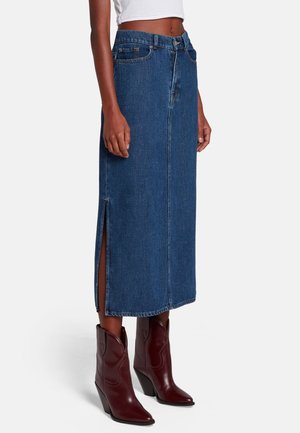 Frau trägt einen hoch taillierten blauen Denim-Midirock mit seitlichem Schlitz und burgunderrote Stiefeletten mit spitzer Spitze und Blockabsätzen.