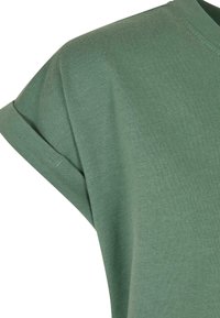 Primo piano di una maglietta verde a maniche corte con risvolto piegato e trama visibile del tessuto a maglia su sfondo bianco.
