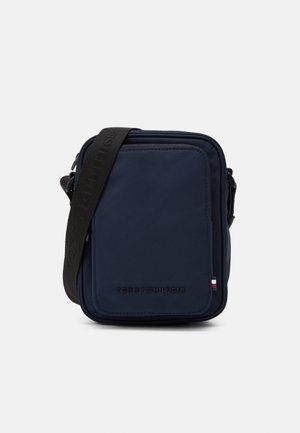Petit sac bandoulière bleu marine avec une bandoulière noire réglable et une poche avant, avec un logo discret et un petit détail de drapeau rouge, blanc et bleu.