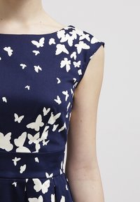 Marinblå klänning med vit fjärilsprint, med en figursydd bodice och korta ärmar. Slät tyg med en lätt glans och plisserad kjol.