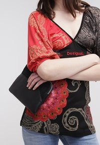 Top de patrón negro y rojo con mangas abullonadas, que presenta intrincados diseños dorados y un acento floral, combinado con un pequeño bolso negro.