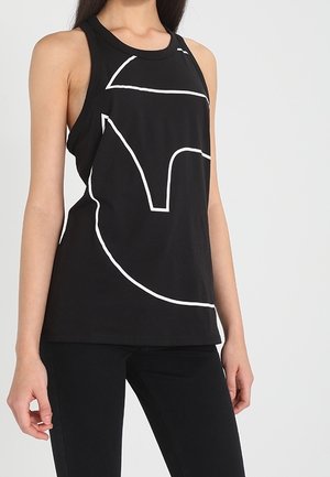 Vrouw draagt een mouwloze zwarte top met een groot wit abstract lijnontwerp, gecombineerd met zwarte broek, staand tegen een effen achtergrond.