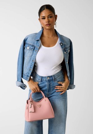 Donna che indossa una giacca di denim blu e jeans sopra una canotta bianca, che tiene in mano davanti una borsa rosa con entrambe le mani.