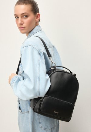 Tânără purtând o geacă ușoară din denim, cu un rucsac negru din piele, prevăzut cu un buzunar frontal cu fermoar și inscripția "Kazar" pe un fundal pal.