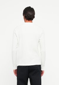 Man met donker haar, gekleed in een effen wit overhemd met lange mouwen en zwarte broek, staand met zijn rug naar de camera tegen een witte achtergrond.