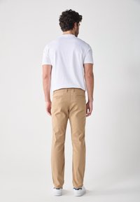 Homme aux cheveux bouclés, portant une chemise blanche à manches courtes, un pantalon beige et des baskets blanches, debout de dos sur un fond uni.