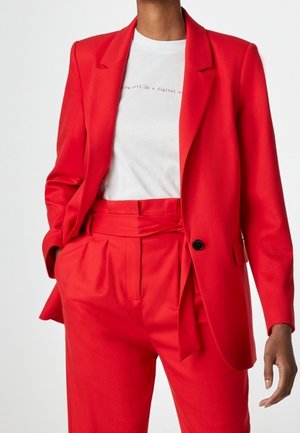 Persoon draagt een opvallend rood pak met een blazer met één knoop en een broek met hoge taille, gecombineerd met een wit T-shirt met afbeelding.