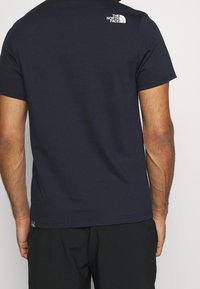 Camiseta de algodón azul oscuro con mangas cortas y cuello redondo. Presenta un logo blanco en la parte superior de la espalda y una pequeña etiqueta en el dobladillo.