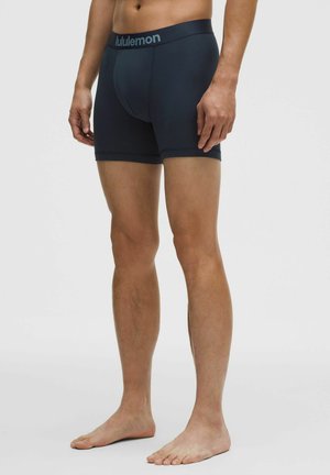 Homme portant un boxer Lululemon noir, debout pieds nus contre un fond clair uni, montrant une vue de face et de côté.