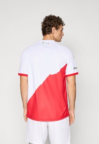 Rot-weißes Sportshirt mit kurzen Ärmeln und einem diagonalen Farbblockdesign, das eine glatte Textur aufweist und ein kleines Logo am Ärmel hat.