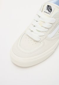 Ljusbeige mockaskor med vita snören, perforerat tåparti och ljusblå detaljer. Utrustad med klassisk Vans-logotyp på tungan.