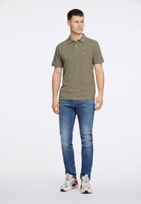 Olijfgroene polo met een kraag en korte mouwen, gecombineerd met blauwe jeans en crème sneakers met rode accenten. Eenvoudig, aansluitend ontwerp.