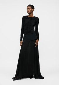 Žena stojící, oblečená v dlouhém černém topu s řasením a dlouhými rukávy a v černé dlouhé sukni s černými špičatými botami.