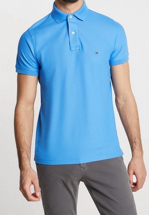 Mand iført en lyseblå kortærmet poloshirt med lille logo på brystet og grå bukser, stående med afslappede arme.