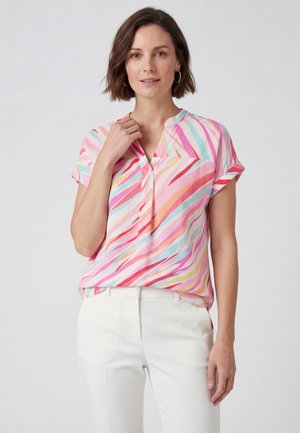 Femme portant un chemisier rose à rayures multicolores manches courtes et un pantalon blanc, debout devant un fond clair uni.