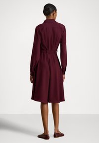 Bordové šaty s dlhými rukávmi, priliehavým pásom a rozšírenou sukňou. Vyrobené z látky s hladkým povrchom, obsahujú golier a detail na zaväzovanie.