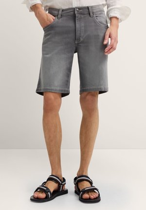 Mann trägt graue Jeansshorts und schwarze Sandalen, steht mit einer Hand in der Hosentasche vor einem einfarbigen Hintergrund.