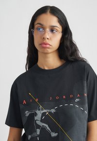 Νέο άτομο με μακριά σκούρα μαλλιά και σκουλαρίκι στη μύτη, φορώντας γυαλιά χωρίς σκελετό και μαύρο μπλουζάκι Air Jordan με γραφικά, σε απλό φόντο.