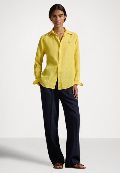 Sieviete stāv, valkājot dzeltenu pogājamu kreklu, vaļīgas melnas bikses un brūnas sandales uz vienkārša balta fona.