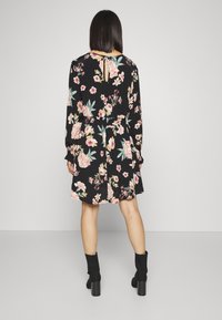 Kvinna stående med ryggen mot kameran, iförd en svart blommig klänning med långa ärmar och svarta klackiga ankelskor mot en enfärgad bakgrund.