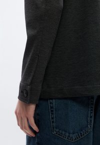 Σκούρο γκρι πουκάμισο με υφή και μανσέτα με κουμπί. Συνδυάζεται με μπλε τζιν παντελόνι που εμφανίζει λεπτομέρειες στις τσέπες.