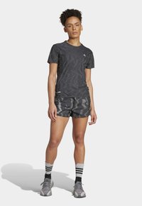 Graues Tarn-T-Shirt mit dezentem Wellenmuster, kombiniert mit passenden Shorts, die sich wiederholende geometrische Designs aufweisen. Sportliche Schuhe und gestreifte Socken.