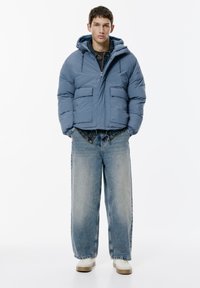 Ung man i en blå vadderad huva-jacka, denimskjorta, vida blekta jeans och vita sneakers, står mot en vit bakgrund.