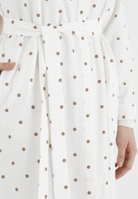 Robe blanche avec une ceinture, ornée d'un motif de pois bruns. Le tissu semble lisse et léger, avec des manches longues.