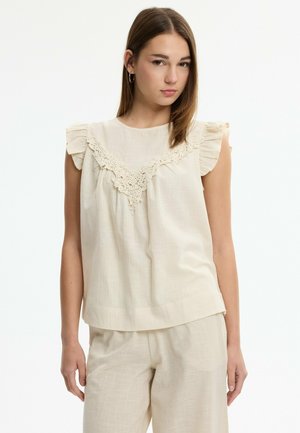 Joven mujer con top beige sin mangas con volantes en los hombros y detalles de encaje, combinada con pantalones sueltos a juego, de pie frente a fondo blanco.