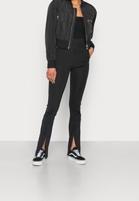 Veste bombardière noire avec fermeture éclair, associée à un pantalon noir ajusté avec des fentes aux chevilles et des baskets noires et blanches.
