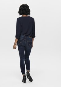 Jean skinny blu scuro con tasche posteriori caratterizzate da un motivo di cucitura dettagliato, abbinati a una camicia nera con bottoni e scarpe con tacco nere.