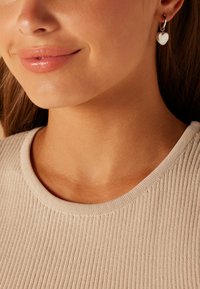 Zilveren oorringen met hartvormige pendants, die het licht weerkaatsen. Het model draagt een ribbelige beige top met een ronde hals.