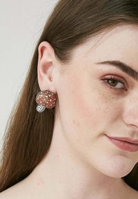 Runda örhängen med orange och klara rhinestones, silverdetaljer och en texturerad yta, bärs i modellens öra.