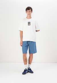 White cotton t-shirt with black Adidas logo patch, paired with blue denim shorts. Model wears blue sneakers with three white stripes.