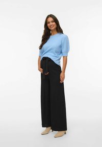 Modra pletena jopica s kratkimi puff rokavi; črne široke hlače z vezalko; svetlo bež čevlji na peto. Enobarvna ozadje.