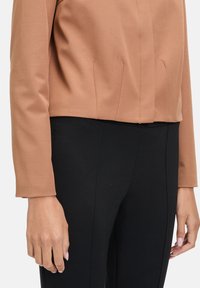 Braune, taillierte Jacke aus strukturiertem Stoff, mit langen Ärmeln und sauberen Nähten, kombiniert mit schwarze, taillierte Hose. Einfaches Design, ohne Muster oder Hardware.