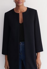 Schwarzer Wollmischmantel mit geradem Schnitt, Rundhalsausschnitt und 3/4-Ärmeln. Verfügt über eine saubere Vorderseite ohne Knöpfe, kombiniert mit einem schwarzen Oberteil.