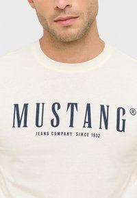 Kremowa koszulka z krótkim rękawem wykonana z bawełny, z napisem "MUSTANG" w pogrubionym granatowym kolorze i okrągłym dekoltem.