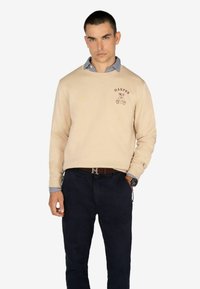 Sudadera beige con diseño de oso de peluche bordado y texto "HARPER", combinada con pantalones oscuros y un cuello de camisa a cuadros.